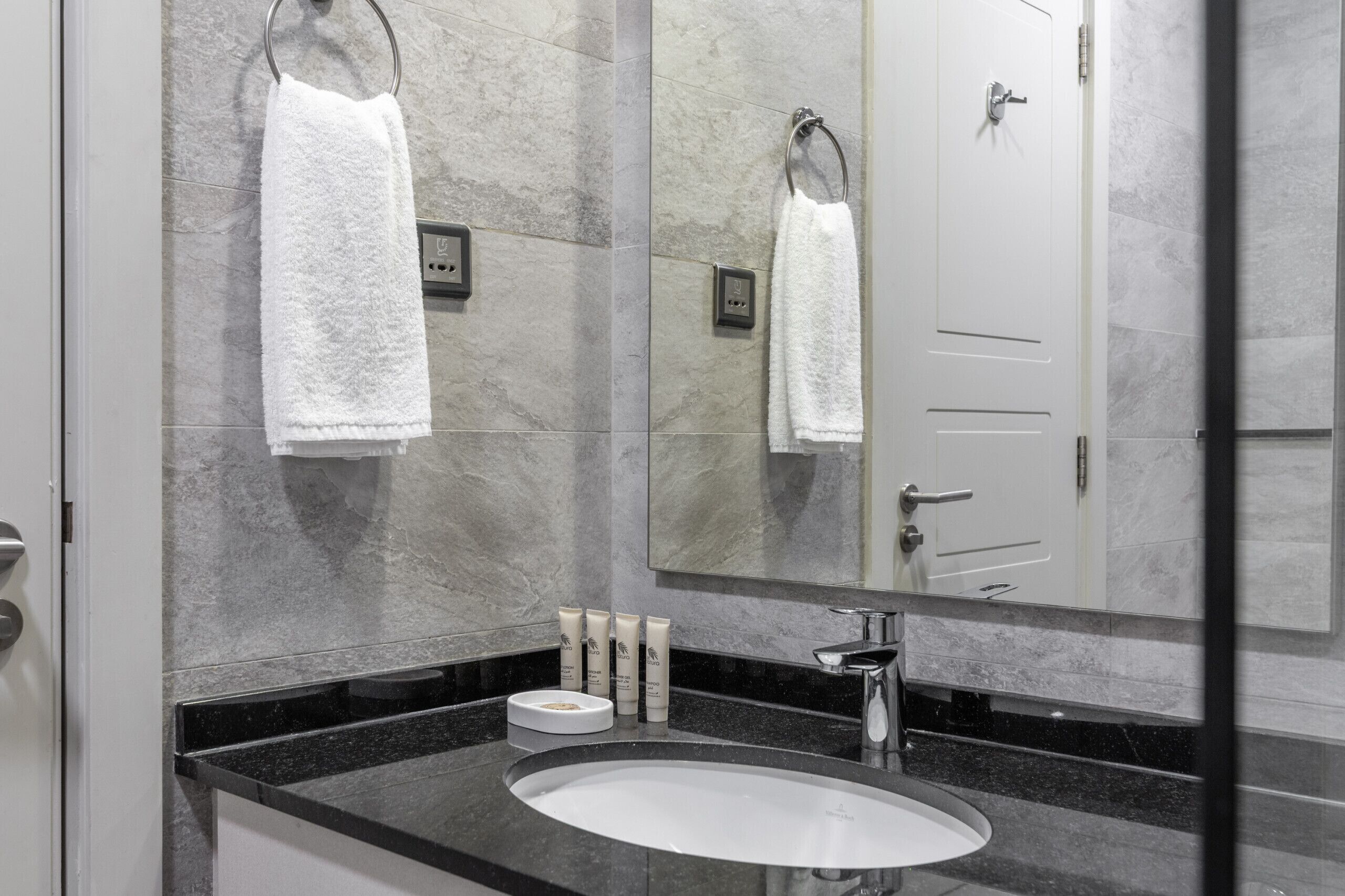 Une salle de bain avec un mur en marbre, un lavabo blanc et un grand miroir qui reflète la porte et les serviettes.
