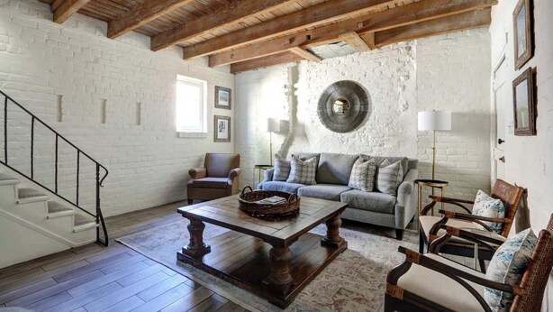 A spacious living room with a wooden ceiling, white brick walls, a grey sofa, a coffee table, and a staircase. - The Gaston Loft, Historic | Steps to Forsyth Park (Savannah)