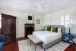 A bedroom with a bed, a bench, a dresser, a nightstand, a lamp, a ceiling fan, and a fireplace. - The Gaston Loft, Historic | Steps to Forsyth Park (Savannah)