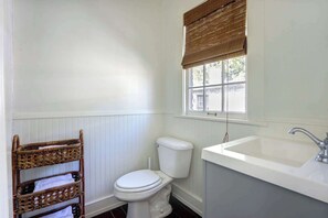 A bathroom with a white toilet, a sink, and a window with blinds. - The Gaston Loft, Historic | Steps to Forsyth Park (Savannah)