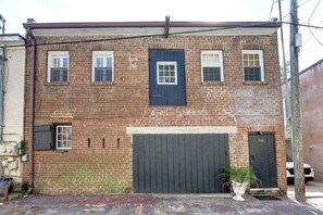 A two-story brick building with a dark blue door and windows, a garage door, and a small planter with a palm plant. - The Gaston Loft, Historic | Steps to Forsyth Park (Savannah)