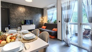 Ein modernes Wohnzimmer mit einer Couch, einem Esstisch für zwei Personen, einem Fernseher und einem Balkon mit Vorhängen.