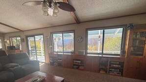 Una sala de estar con sofá, mesa de centro y una estantería llena de libros. Amplias ventanas ofrecen vistas al exterior.