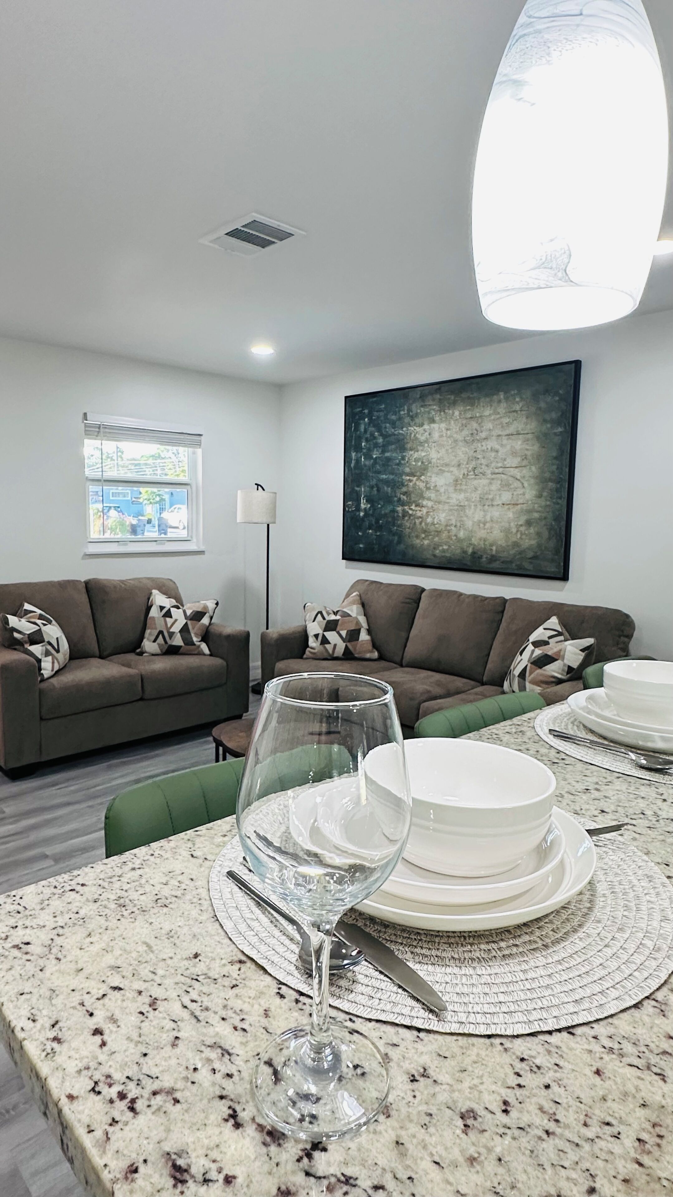 A modern living room with a dining area, featuring a sofa set, a glass table with plates and a wine glass, and a large abstract painting on the wall.