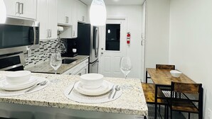 Een moderne keuken met witte kasten, een granieten aanrecht, een ingebouwde magnetron en een roestvrijstalen koelkast. Er is een eethoek met een houten tafel en stoelen, en een witte deur op de achtergrond.