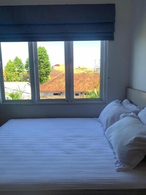 A bedroom with a bed, white bedding, and a view of buildings and trees through a window with a dark valance. - Sanur House Lavender (Denpasar)