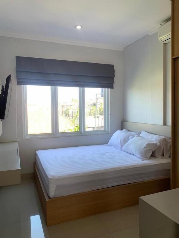 A bedroom with a bed, a window with a view, a bedside table, and a wall-mounted air conditioning unit. - Sanur House Lavender (Denpasar)