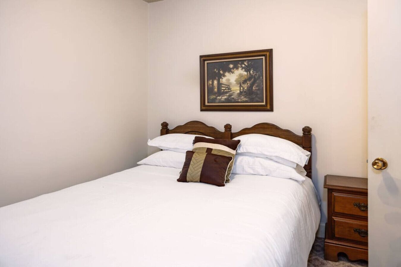 A neatly made bed with white linens and a brown pillow, a wooden headboard, and a small wooden nightstand with drawers.