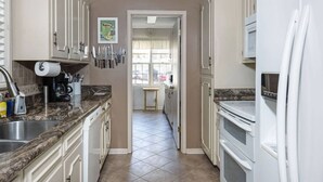 Une cuisine avec des meubles blancs, un plan de travail en marbre et un réfrigérateur. Il y a une fenêtre avec des rideaux et une porte qui donne sur une autre pièce.