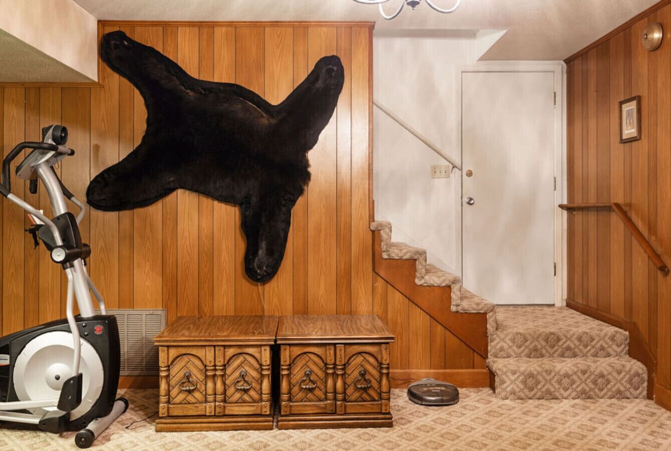 Ein Zimmer mit einem aufgehängten Tierkopf, einem Laufband, einer Holzkommode und einer Treppe.