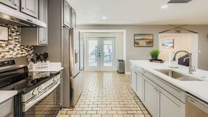 A modern kitchen with a stainless steel refrigerator, a built-in oven, and a dishwasher. There is a white countertop with a sink and a potted plant. The floor is tiled, and the kitchen has a view of the outside through a glass door. - New Sunrise Sanctuary, 4BR home w/ pool & jacuzzi (Tampa)