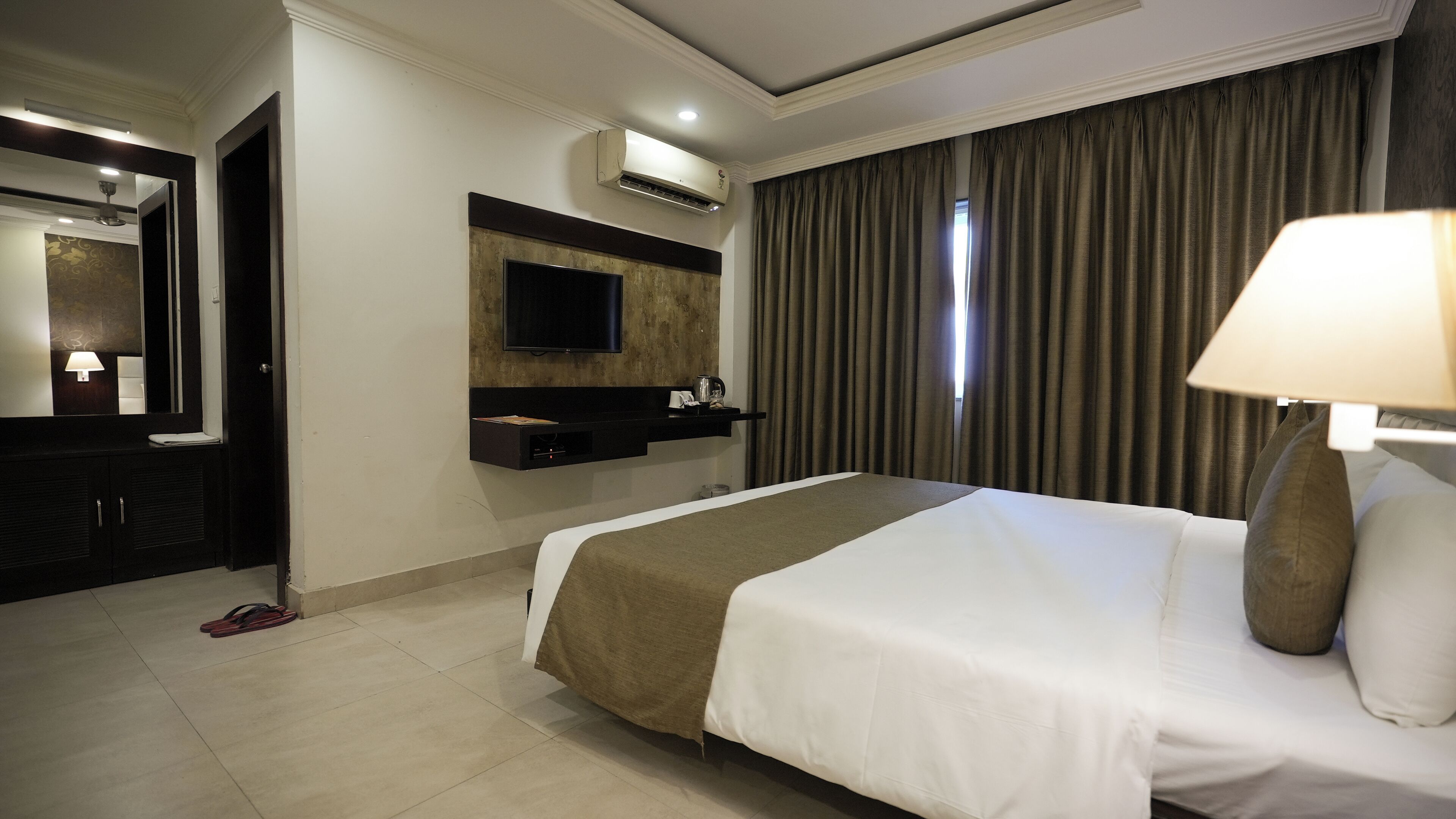 A hotel room with a large bed, a flat-screen TV mounted on the wall, a minibar, and a window with curtains.