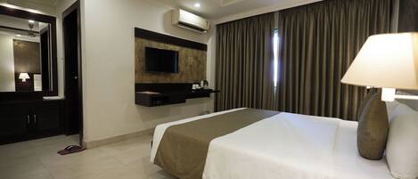 A hotel room with a large bed, a flat-screen TV mounted on the wall, a minibar, and a window with curtains.