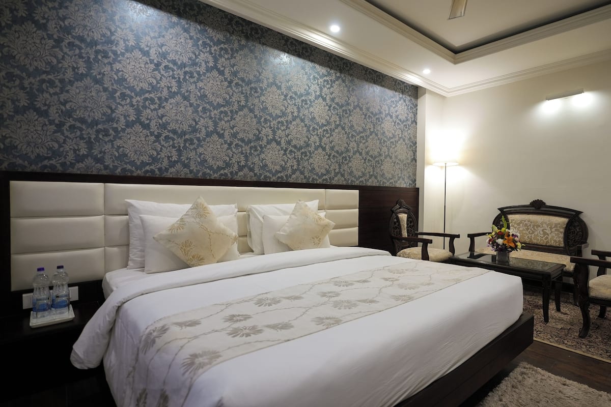 A hotel room with a large bed, a wooden chair, a small table with a vase of flowers, and a wall with a blue patterned wallpaper.