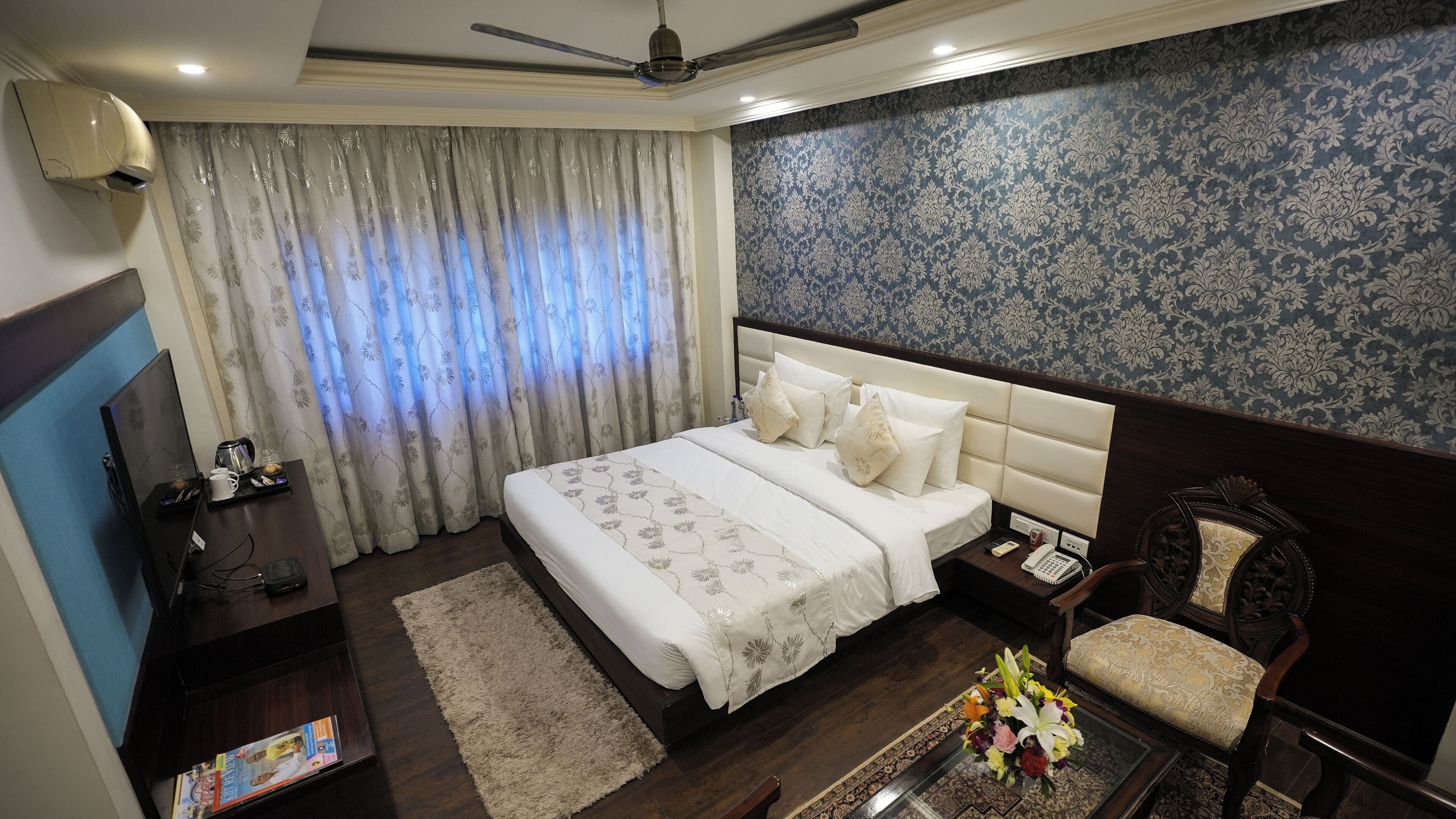 A hotel room with a large bed, a desk with a computer, a chair, and a small table with a floral arrangement.