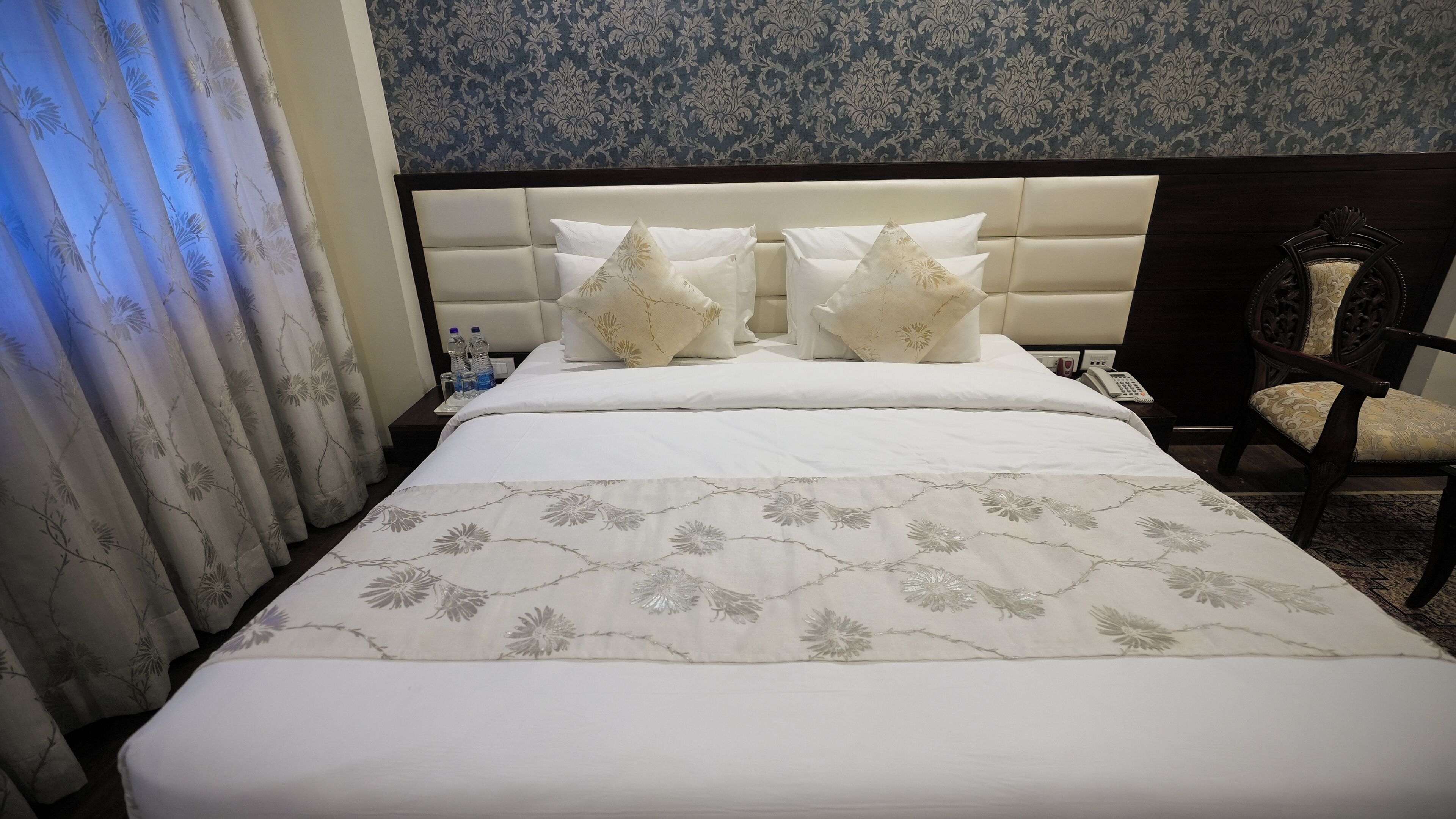 A hotel room with a large bed, two pillows, a bedside table with water bottles, a chair, and a telephone.