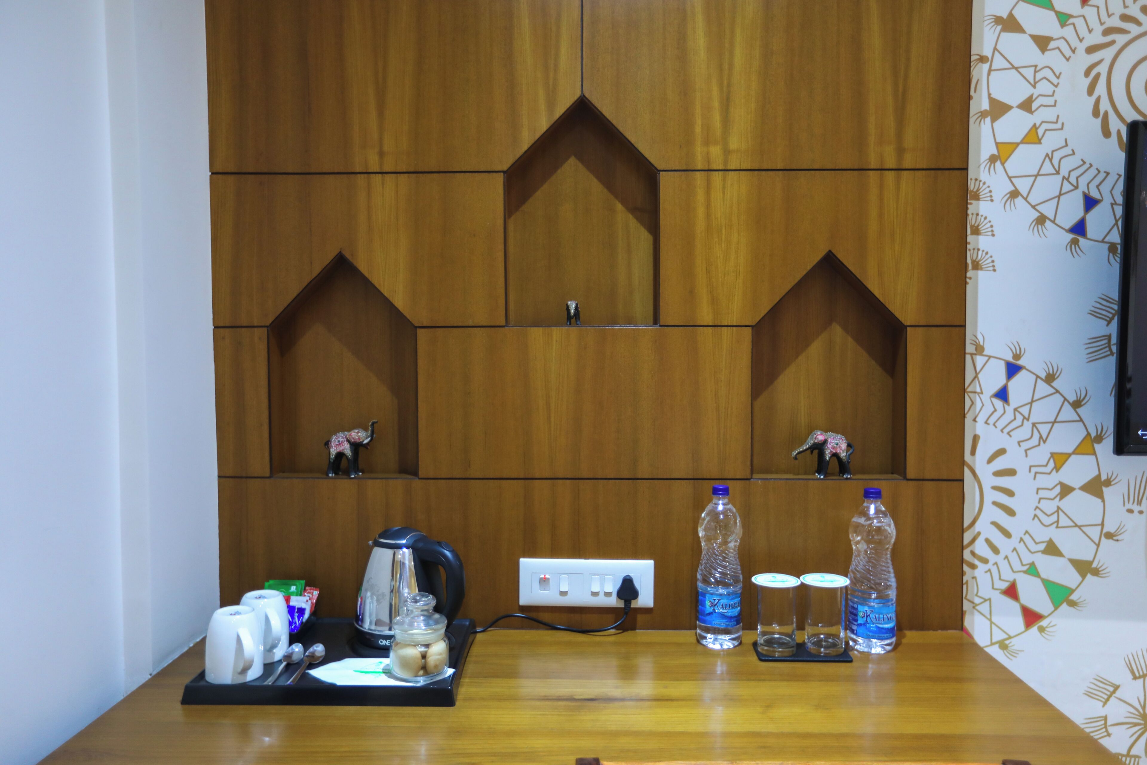 A wooden wall with geometric cutouts, a power outlet, and a table with a coffee maker, cups, a jar, and two bottles of water.