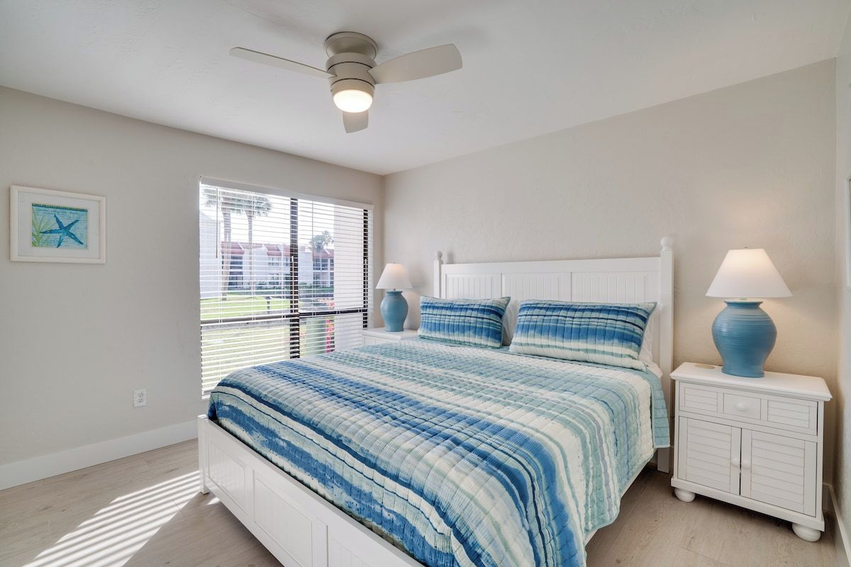 A bedroom with a bed, a ceiling fan, a window with blinds, and a nightstand with a lamp.
