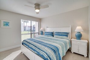 Un dormitorio con cama, ventilador de techo, ventana con persianas y mesita de noche con lámpara.