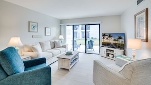 Ein Wohnzimmer mit einem weißen Sofa, einem blauen Sessel und einem Couchtisch. Ein Fernseher steht auf einem Schrank, eine Lampe und ein gerahmtes Bild zieren die Wand.