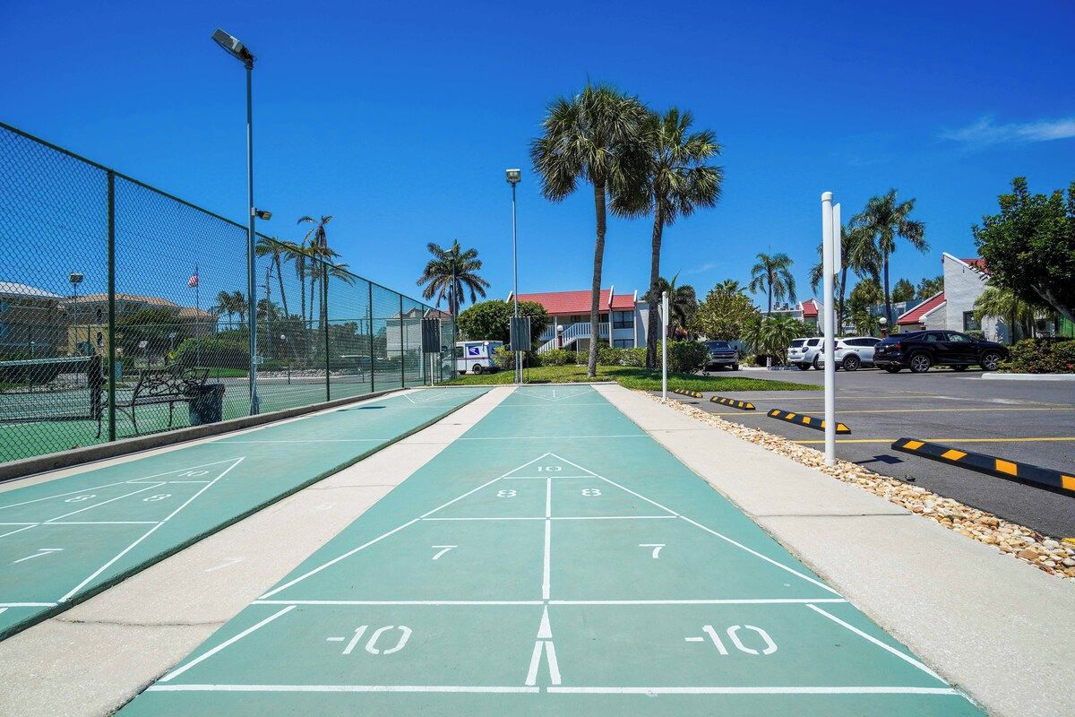 Een jeu de boules baan met een duidelijke weg naar een parkeerplaats, omringd door een hek en palmbomen.