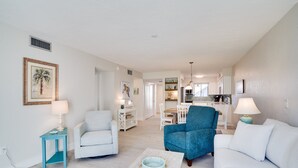 Ein geräumiges Wohnzimmer mit einem hellen Sofa, einem blauen Sessel und einem Couchtisch. Im Hintergrund befindet sich eine Küche mit weißen Schränken und einem Essbereich.