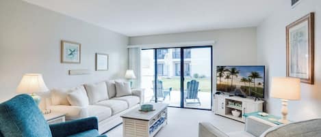 Un salon avec un canapé, une table basse et un téléviseur, un balcon avec des meubles d’extérieur et un tableau encadré au mur.