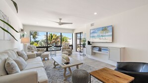 Ein modernes Wohnzimmer mit Sofa, Couchtisch und Hocker, einem Esstisch und einem Gemälde mit Strandmotiv.