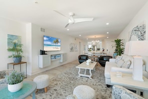 A spacious living area with a ceiling fan, a flat-screen TV mounted on the wall, a dining area with a table and chairs, and a seating area with a sofa and ottomans. - Tropical Sunset 4 (Holmes Beach)