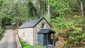 A stone cottage with a small porch, surrounded by lush greenery and a gravel path leading to a road. - Wayside Bothy by Interhome (Drumnadrochit)