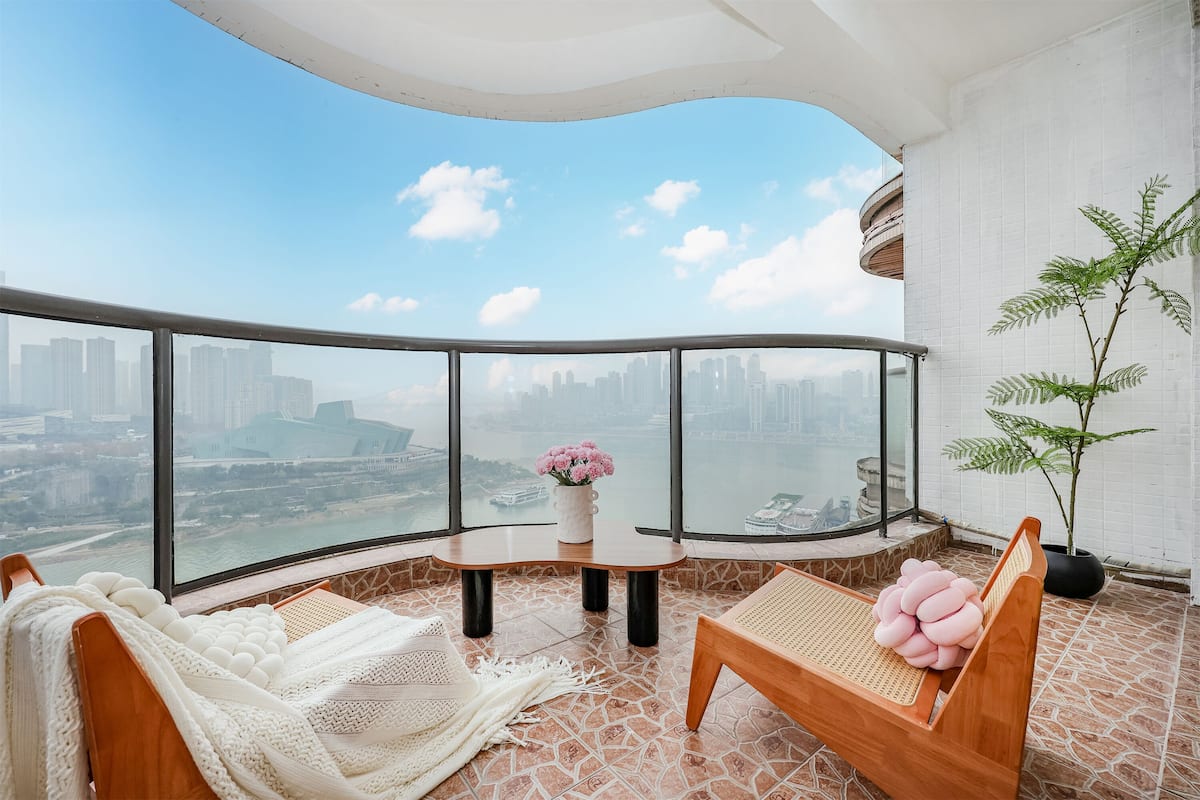 A balcony with a city view, a wooden bench, a cushioned chair, a small table with a vase of flowers, and a potted plant.