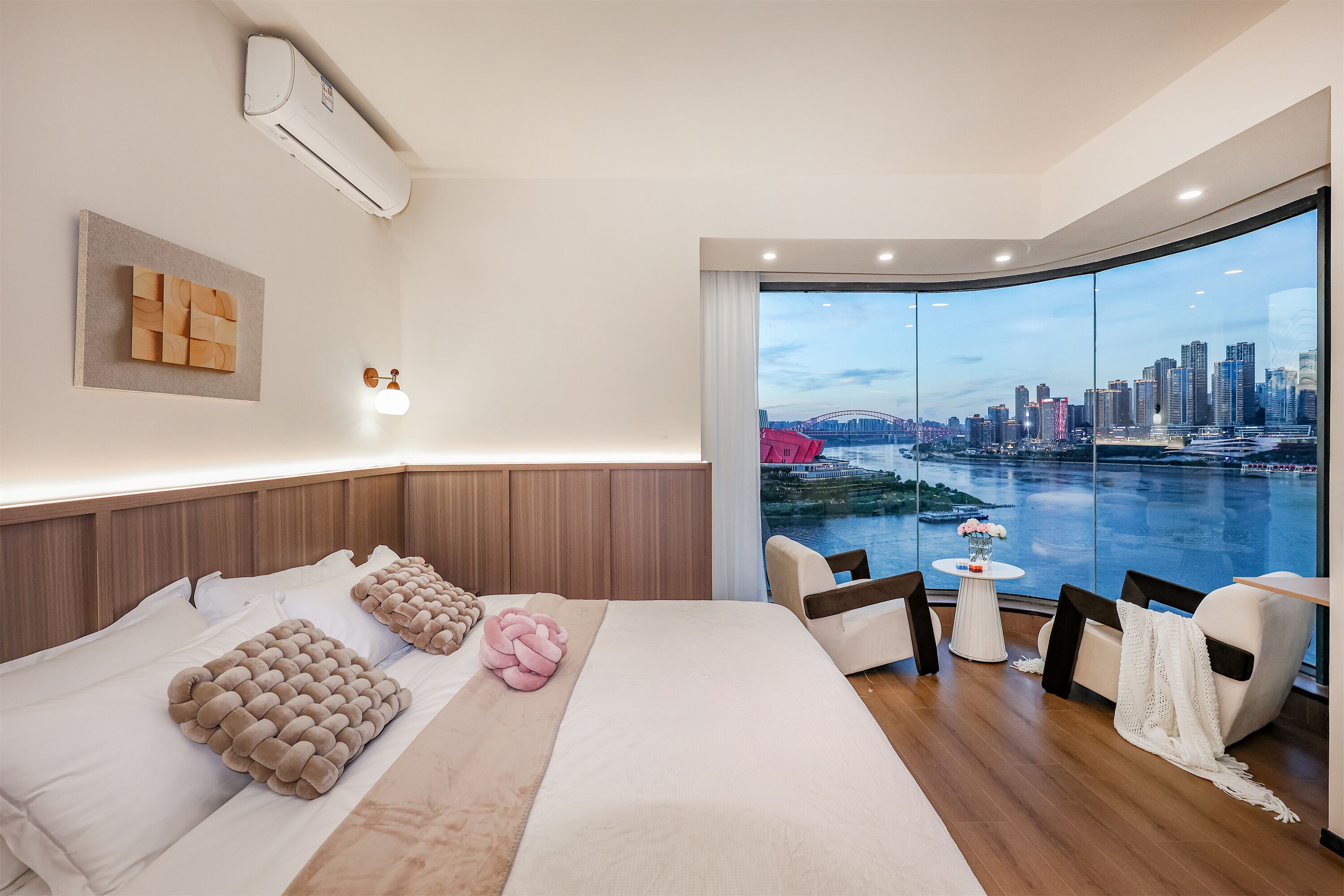 A modern hotel room with a large bed, a view of a cityscape and river, and a seating area with a table and chairs.
