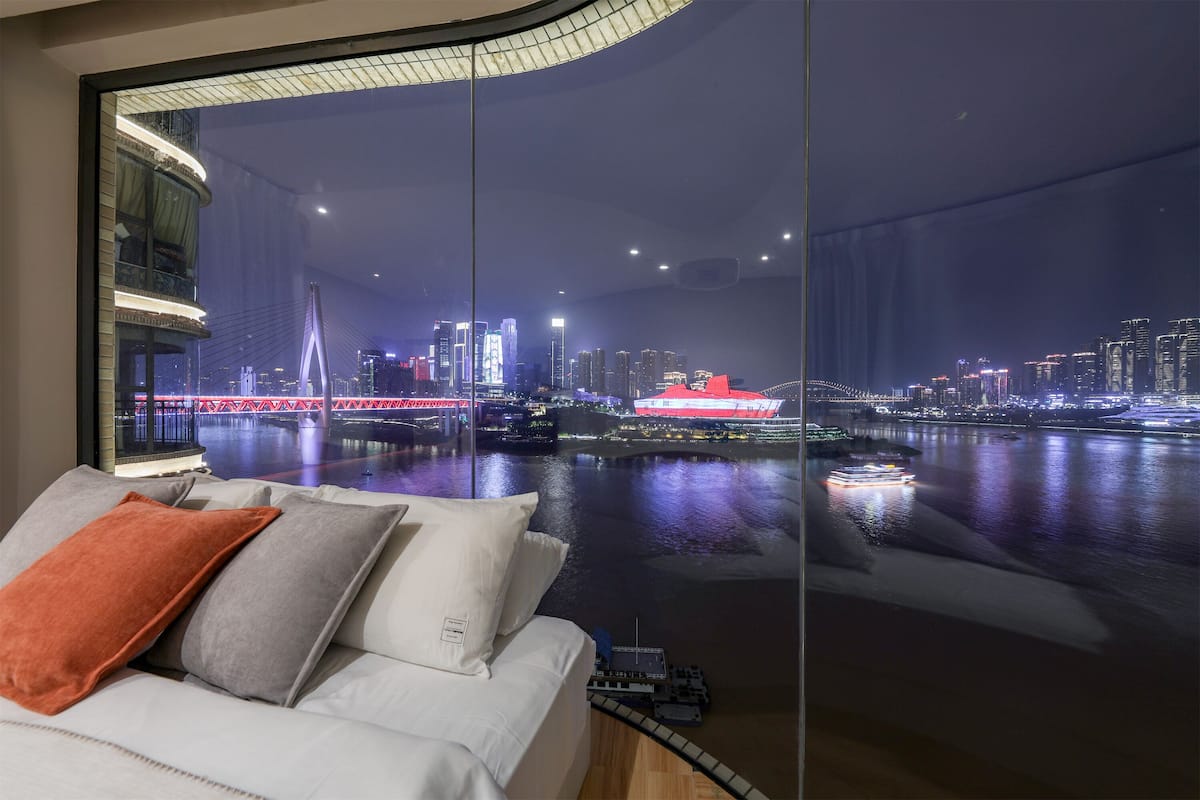 A modern hotel room with a large window overlooking a cityscape at night, featuring illuminated buildings and a bridge.