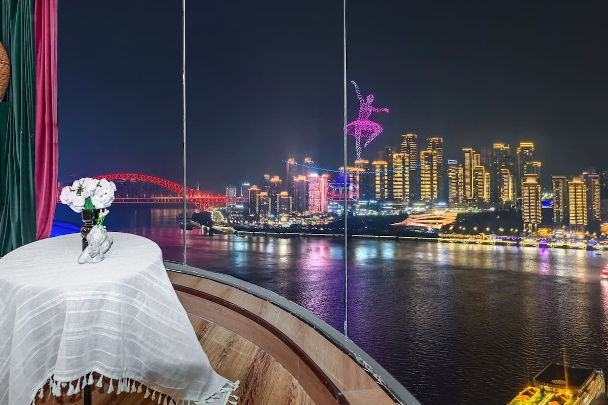 A view from a balcony with a table and vase of flowers, overlooking a cityscape with illuminated buildings and a bridge at night.