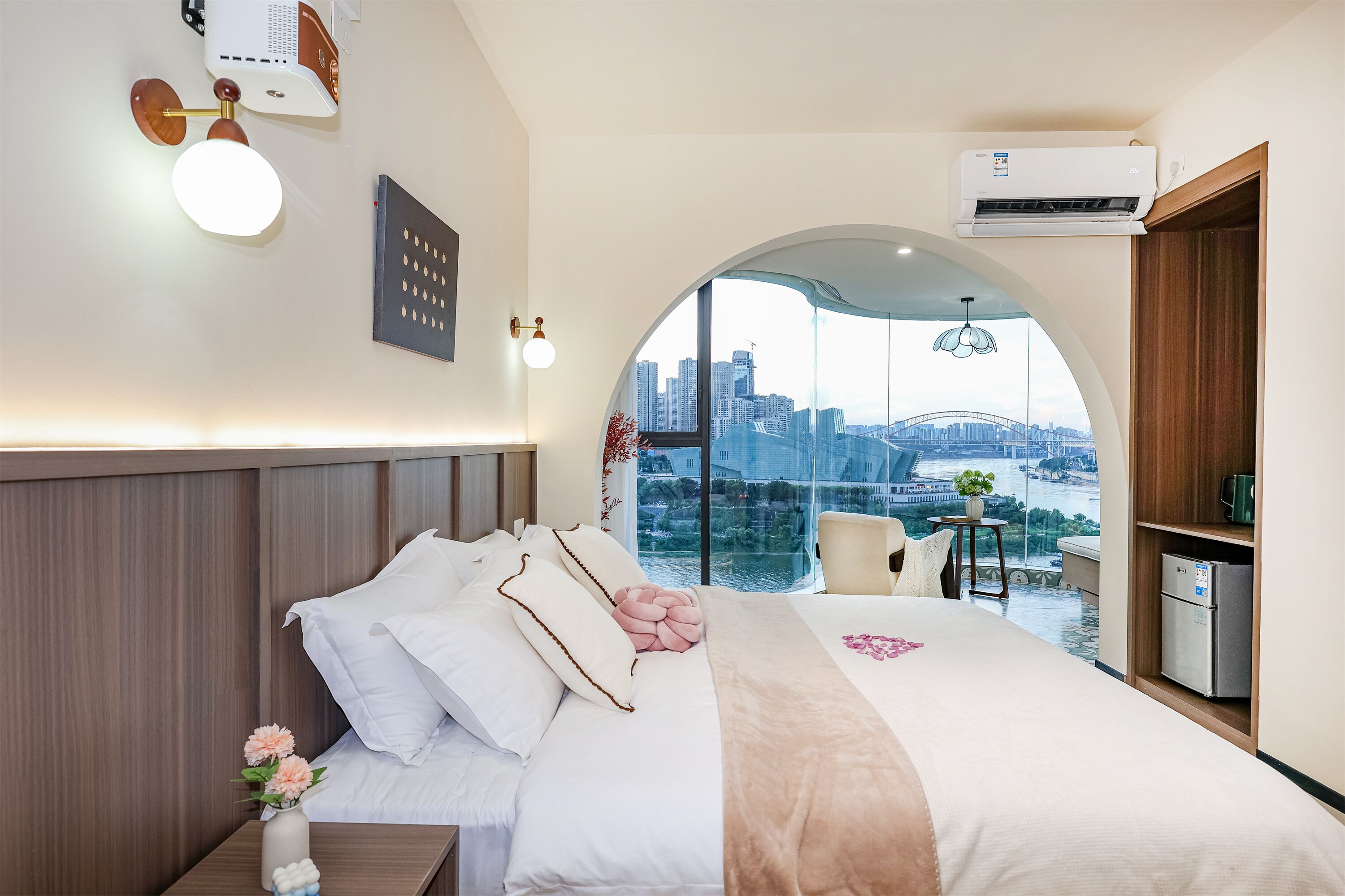 Una camera d'albergo con un letto, un comodino, un piccolo tavolo con una pianta e vista sulla città attraverso una grande finestra.