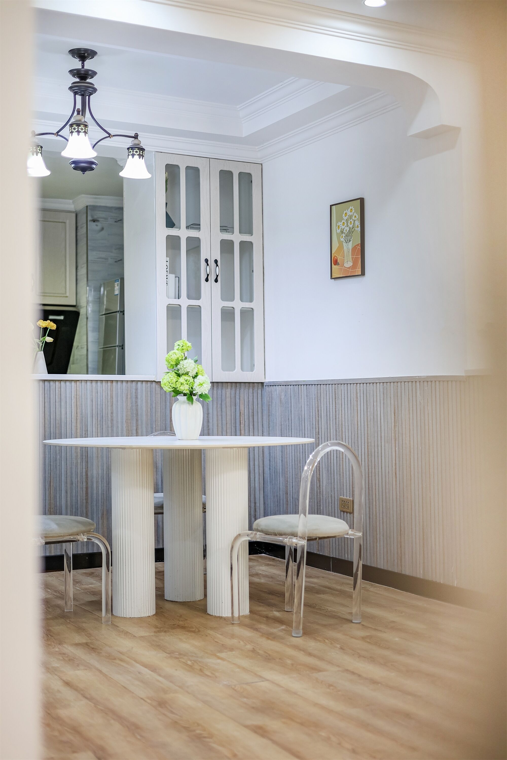 Un coin repas comprenant une table ronde, deux chaises, un vase avec des fleurs, un luminaire au plafond et un meuble avec des portes en verre.
