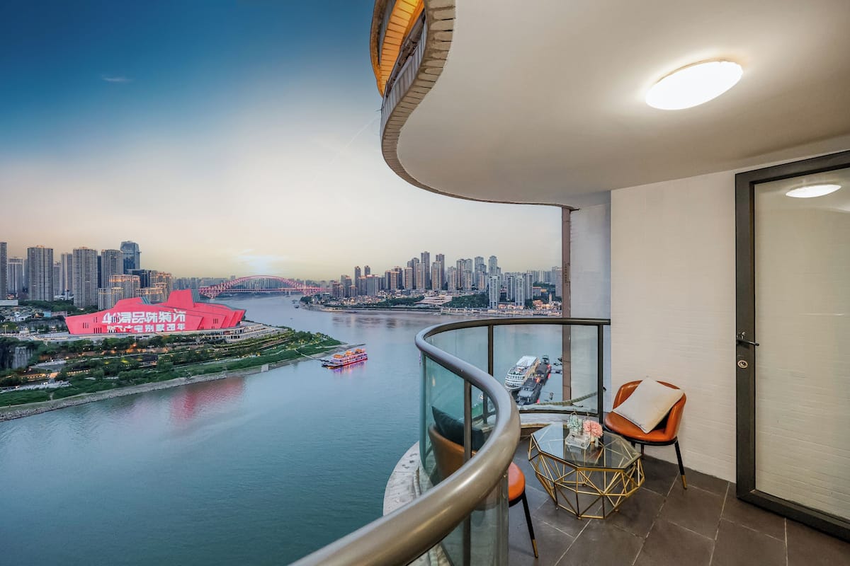 A balcony with a curved railing, a table with chairs, and a view of a cityscape with a red building and a river.