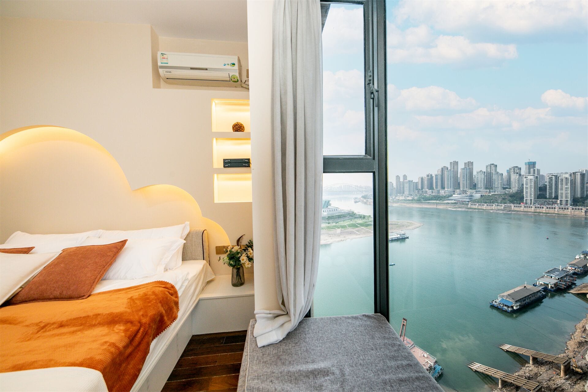 Ein Hotelzimmer mit Bett, einem kleinen Tisch mit einer Vase und Blick auf Stadtansicht und Wasseroberfläche.
