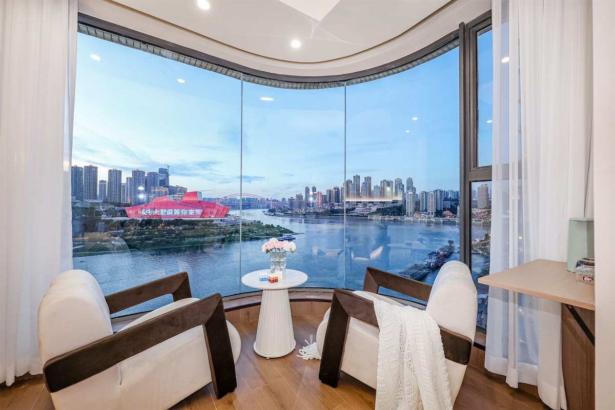 A modern room with a large curved window offering a city view, a small round table with a vase of flowers, and two white chairs.