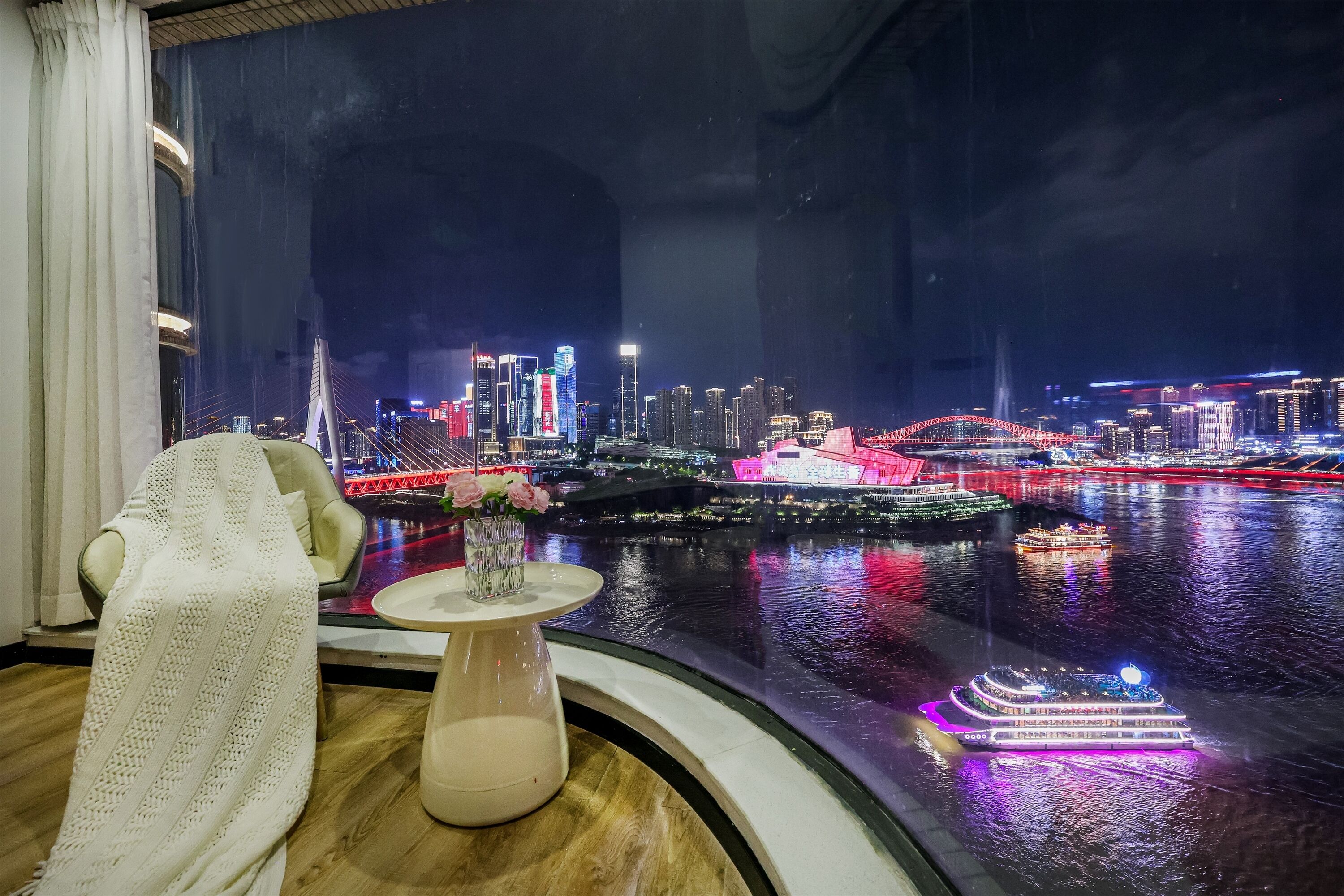 A room with a large window overlooking a cityscape at night, featuring illuminated buildings and a river.