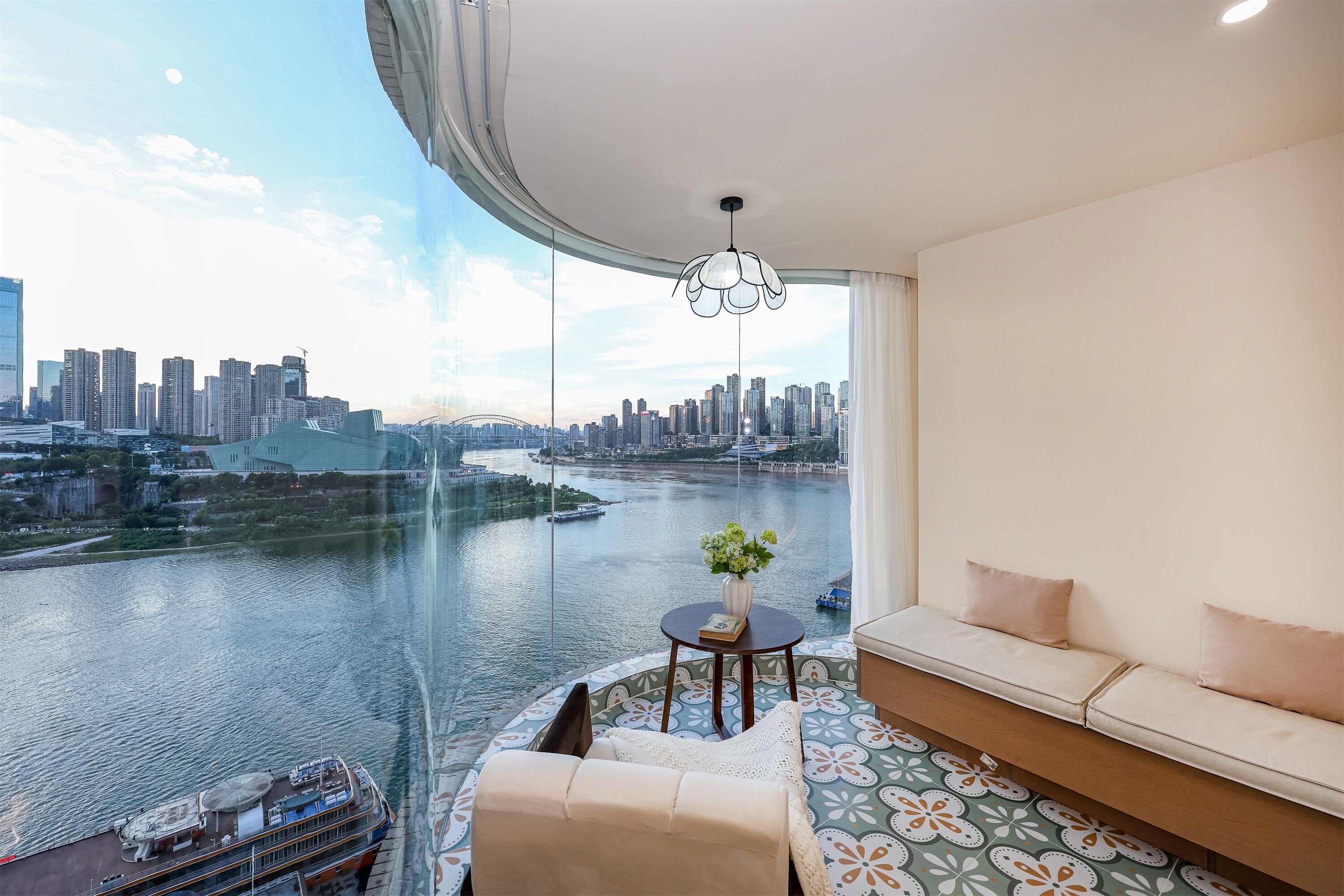 A modern room with a curved ceiling, a sofa, a small table with a vase, and a view of a city skyline and river.