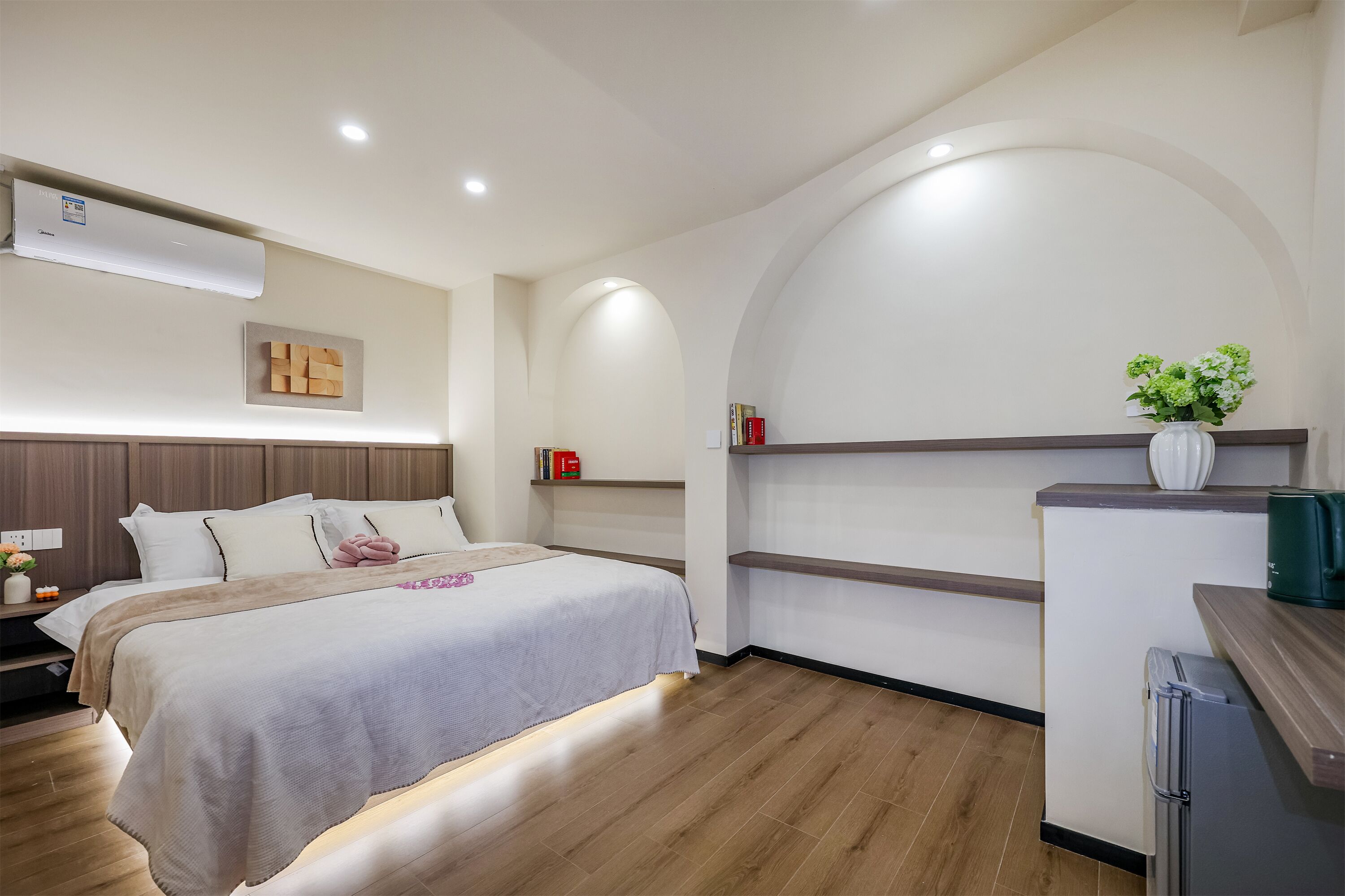 Ein modernes Hotelzimmer mit Bett, Nachttisch, Klimaanlage und einer kleinen Küchenzeile inklusive Kühlschrank und Blumenvase.