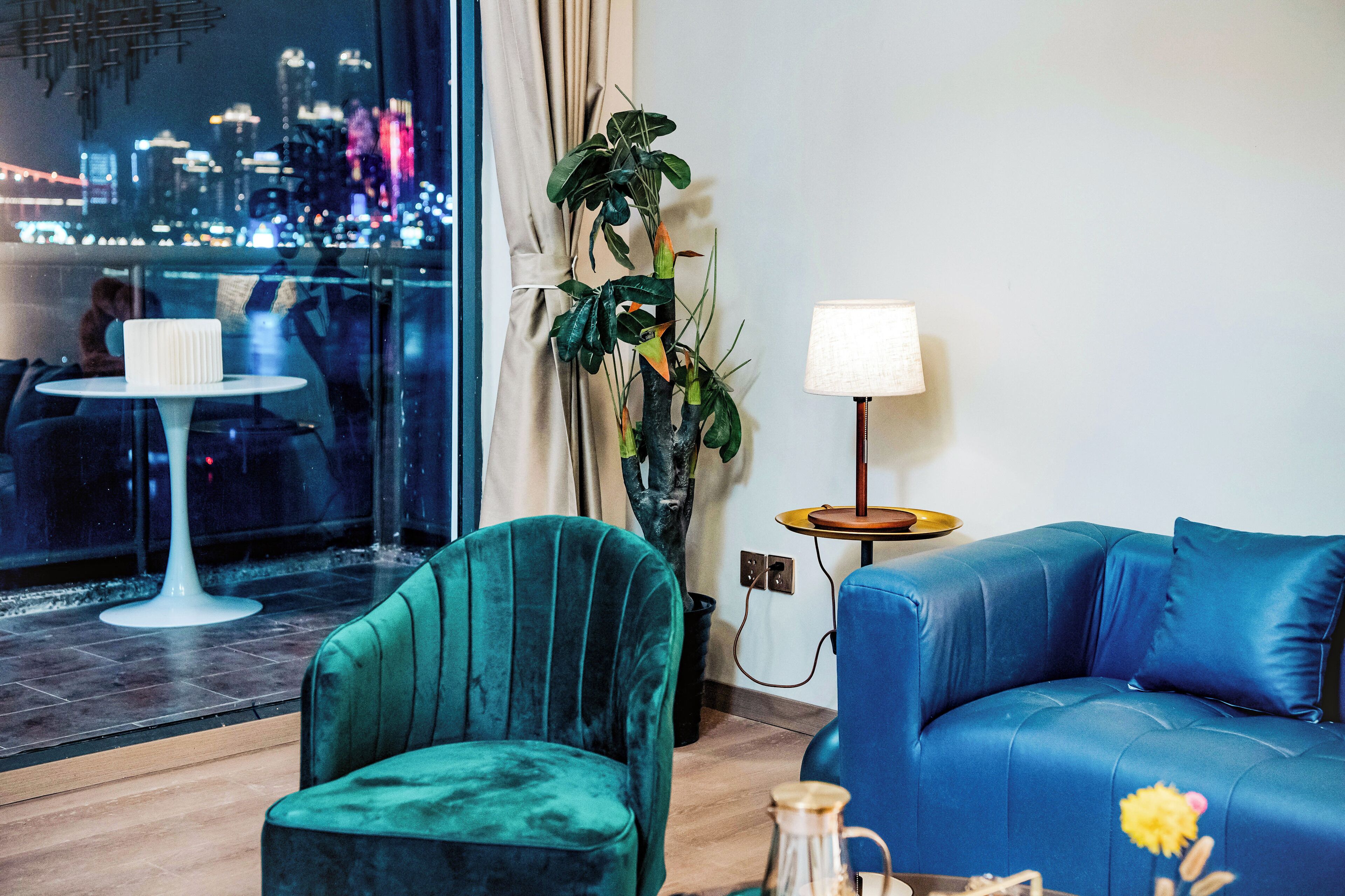 A modern hotel room with a city view, a green armchair, a blue sofa, and a small table with a lamp.