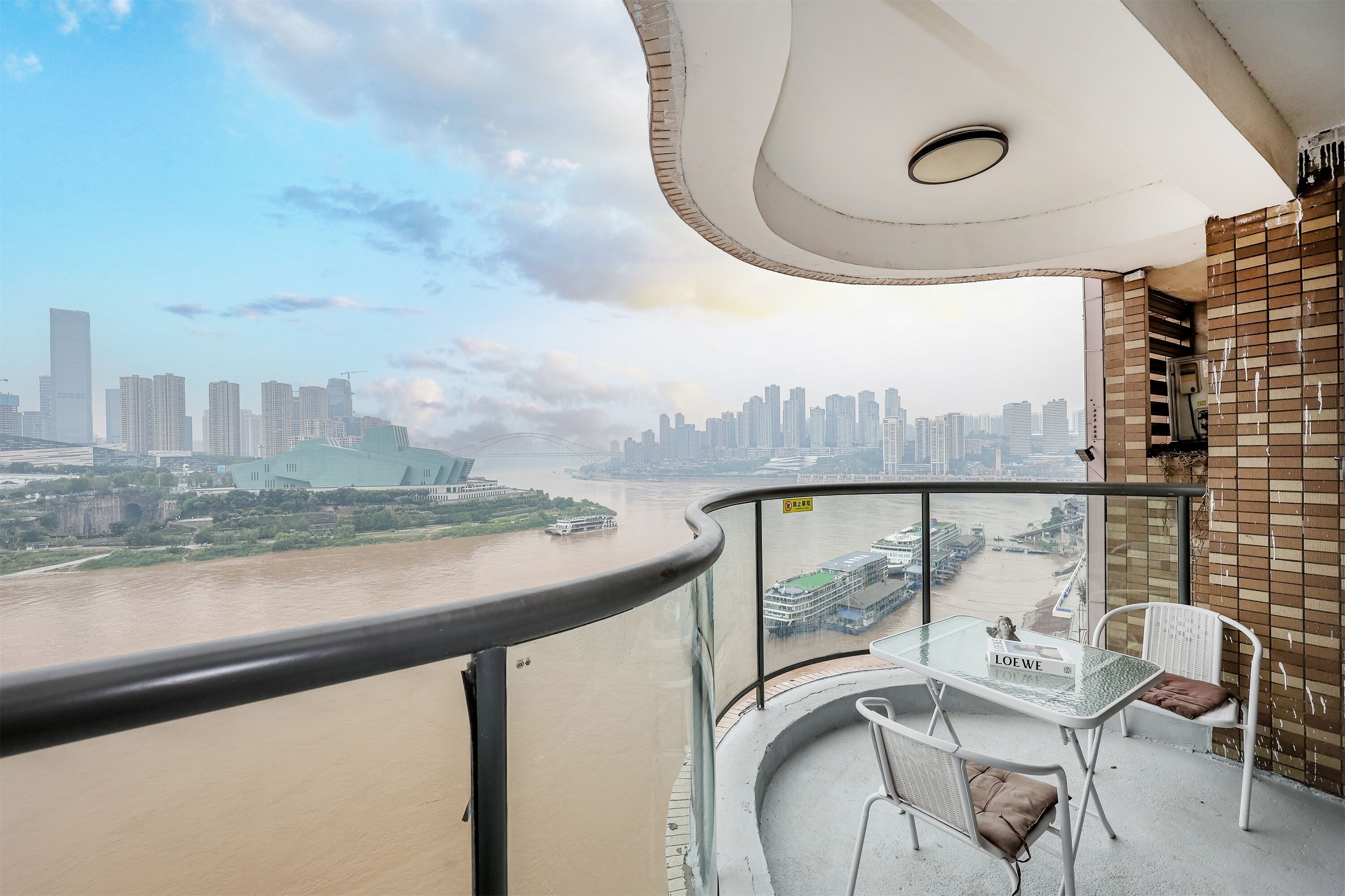 A balcony with a glass table, two chairs, and a view of a river and city skyline.