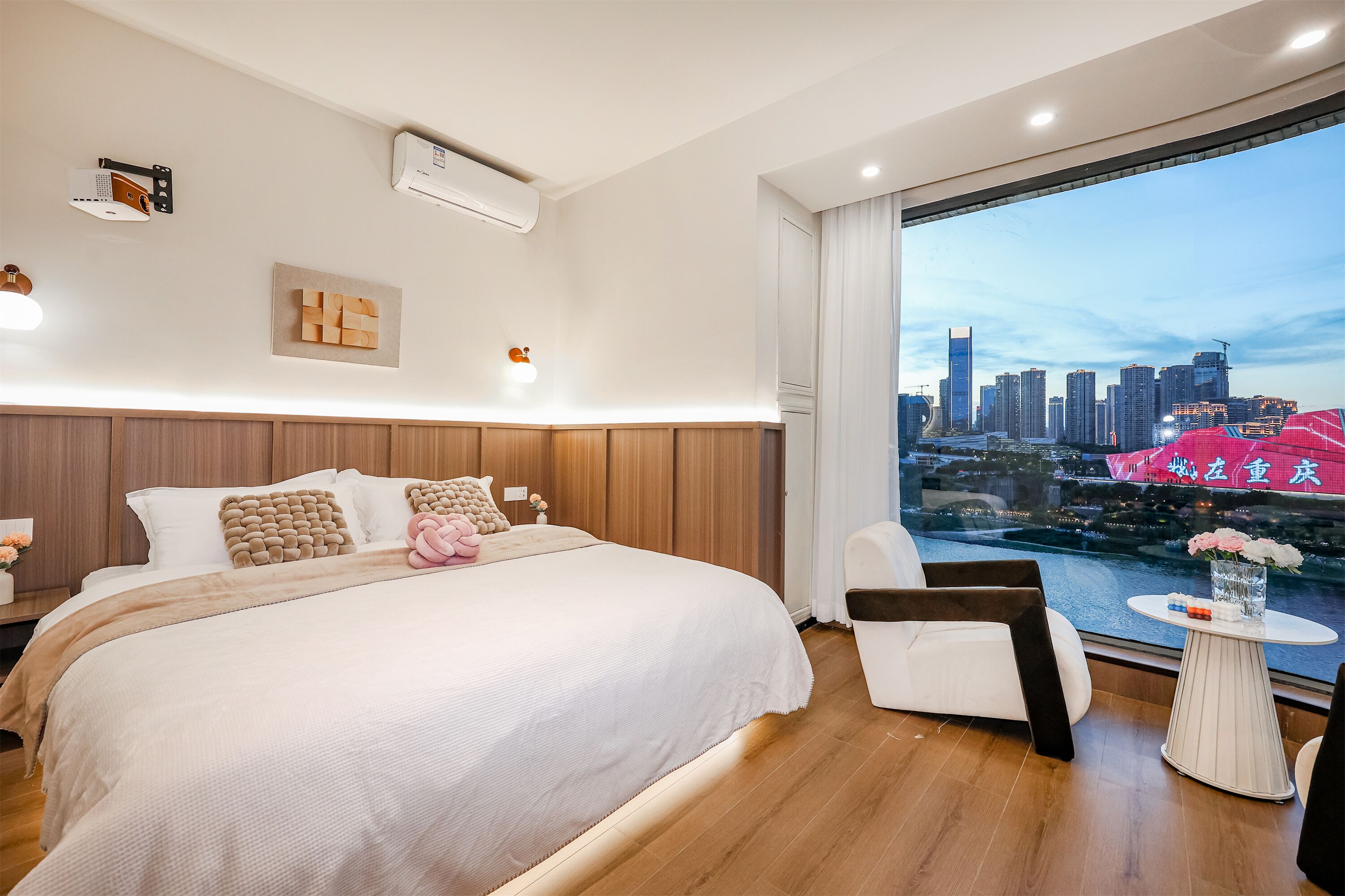 A modern hotel room with a large bed, a city view, and a small table with flowers.