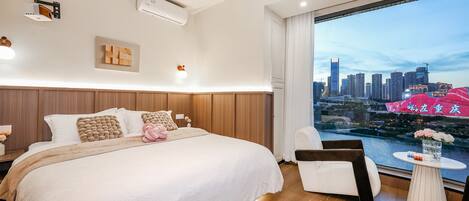A modern hotel room with a large bed, a city view, and a small table with flowers.