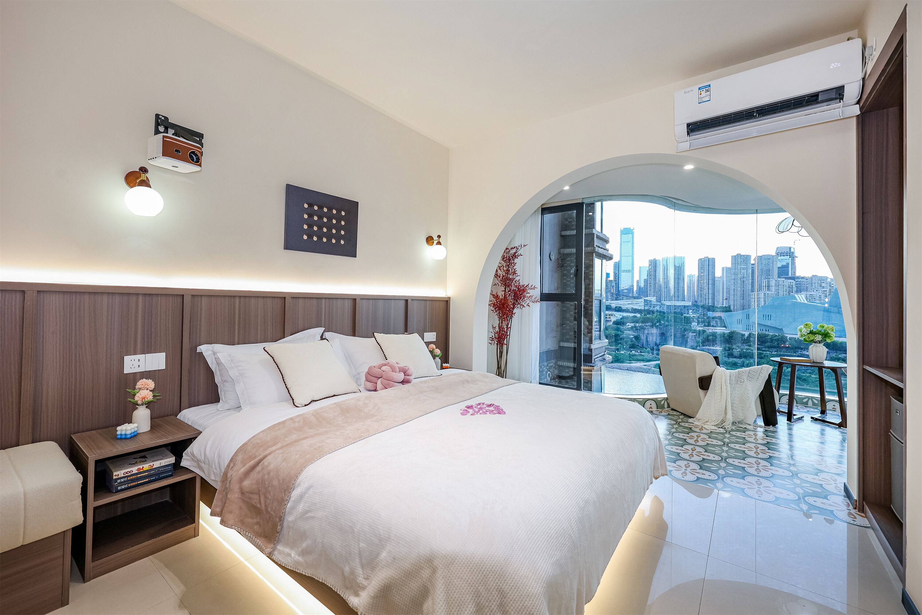 A modern bedroom with a large bed, bedside tables, a nightstand, a small table with a vase, and a view of a cityscape through an arched window.
