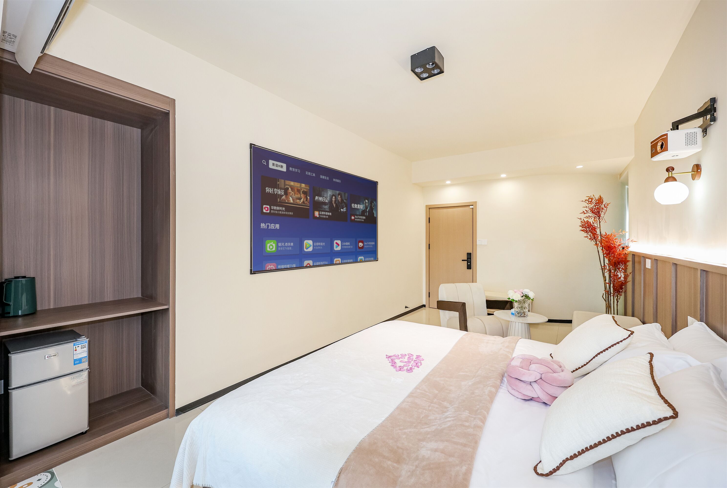 Ein Hotelzimmer mit Bett, kleinem Schreibtisch, Kühlschrank, einem an der Wand montierten Fernseher und einem Holzschrank.