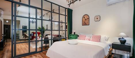 A modern bedroom with a large bed, a chandelier, and a sliding glass door leading to a living area.
