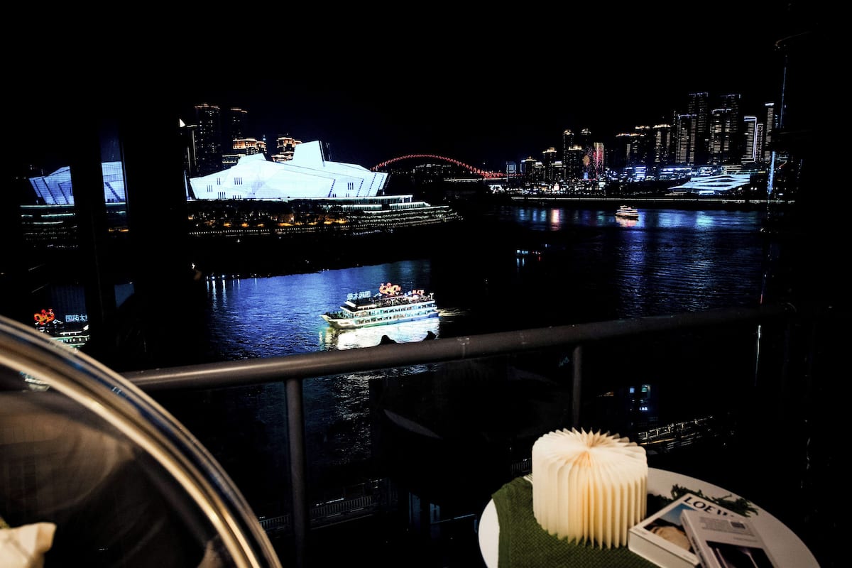 A nighttime view of a city skyline with illuminated buildings and a bridge, a boat on the water, and a dining table with a white napkin and a menu.